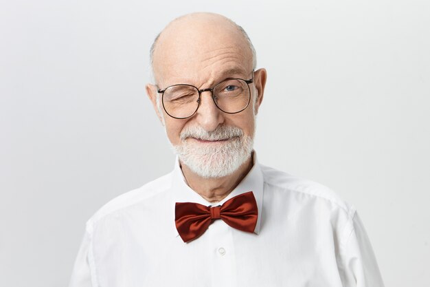 Indoor shot of charismatic charming senior European man wearing elegant red bow tie and eyeglasses having playful facial expression, winking  with smile. Body language and human emotions