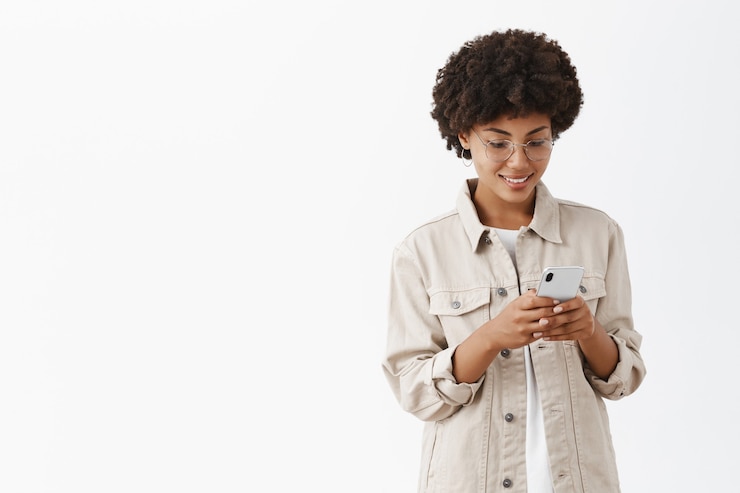 Free Photo indoor shot of charming calm and happy young woman in glasses with afro hairstyle typing message in smartphone gazing with joyful smile at smartphone screen reading interesting article in web