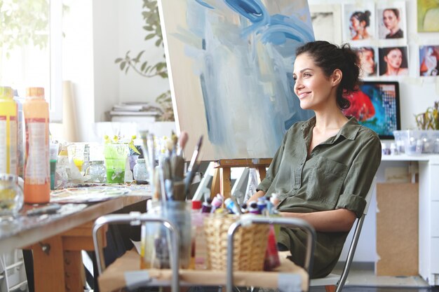 Indoor shot of charming cheerful young European female art teacher with dark curly hair and cute smile sitting at her workshop, surrounded with paints, brushes, waiting for students, looking inspired