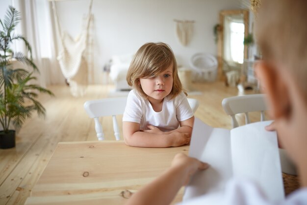 Indoor shot of charming cute little girl sitting at wooden table at home with her unrecognizable young father who is holding sheet of paper, checking her homework. Selective focus on kid's face