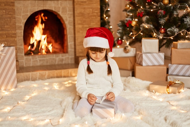 Indoor shot of charming female child wearing white sweater and santa claus hat, opening present box from Santa Claus, posing in festive room with fireplace and Xmas tree.