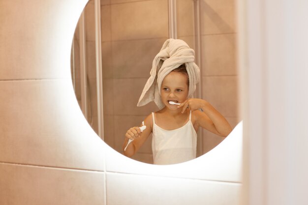 거울 앞에 서서 튜브에서 치약을 짜내며 행복한 미소를 짓고 있는 매력적인 어린 소녀가 욕실에서 이를 닦는 실내 사진.