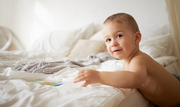 Indoor shot of charming one year old European little child with chubby cheeks standing at edge of bed, trying to climb up, reaching out hand to take book. Bedtime, sleep and childcare concept