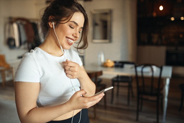 Indoor shot of charming young plus size woman in white top holding smart phone using headset while talking to friend via online video chat, smiling, making plans for weekend. People and technology