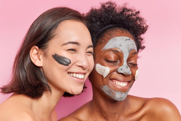 Indoor shot of cheerful beautiful young women stand closely to each other smile gladfully apply clay mask for skin treatment stand topless undergo beauty procedures isolated over pink background