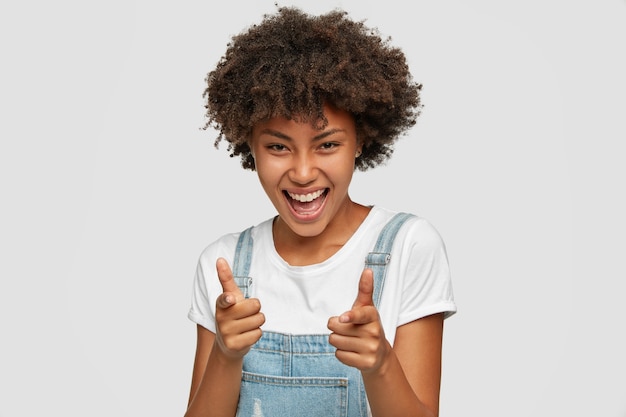 Indoor shot of cheerful carefree ethnic woman points with finger gun gesture  with positive expression