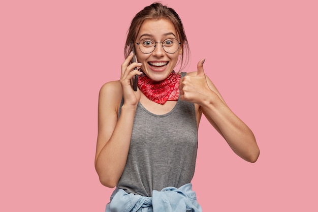 Indoor shot of cheerful Caucasian woman shows okay gesture, keeps thumb raised, expresses her approval, speaks via cell phone