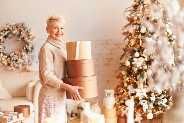 Indoor shot of cheerful elegant middle aged female with blonde short hair standing in decorated living room carrying boxes with gifts, going to hide them until Christmas. Happy New Year concept