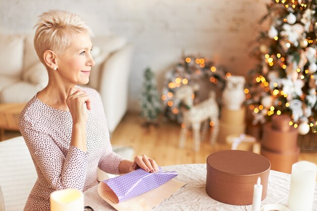 Indoor shot of cheerful mature short haired European female preparing for New Year or Christmas celebration, sitting in living room with gift paper on table, having pensive thoughtful look, smiling