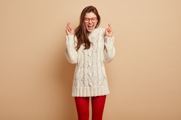 Indoor shot of cheerful overjoyed young female crosses fingers, hopes everything will be fine, wears white jumper, red leggings, has desire for dreams come true, stands against beige wall