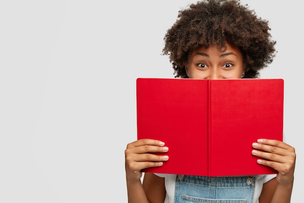 Indoor shot of of cheerful woman covers face with red textbook, has joyful expression