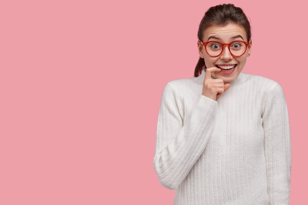 Indoor shot of cheerful young woman keeps index finger near lips, rejoices recieving compliment