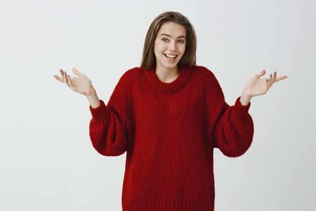 Indoor shot of clueless attractive friendly european woman in trendy red loose sweater, shrugging with spread palms, explaining something, standing unaware and questioned 