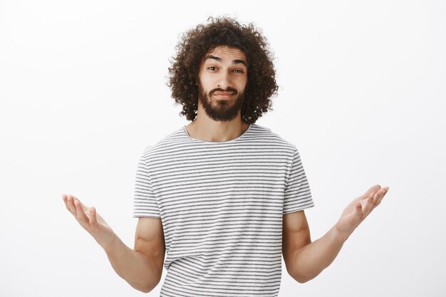 Indoor shot of clueless confused hispanic boyfriend with afro hairstyle and masculine beard, raising palms cluelessly and lifting eyebrows