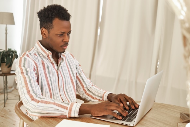 Indoor shot of concentrated African student learning online via laptop using wireless internet connection, sitting at desk, doing homework or preparing for exam.