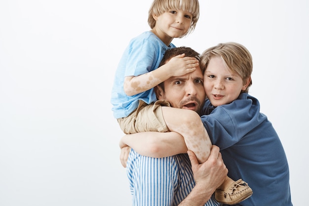 Снимок в помещении: обеспокоенный усталый отец держит симпатичного белокурого сына с витилиго на плечах, хмурится и волнуется, а старший брат висит на груди отца