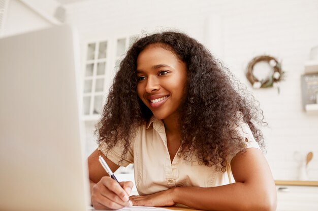 Indoor shot of confident cheerful young dark skinned female freelancer sitting at dining table, working remotely using portable computer. Modern electronic gadgets, job and occupation concept