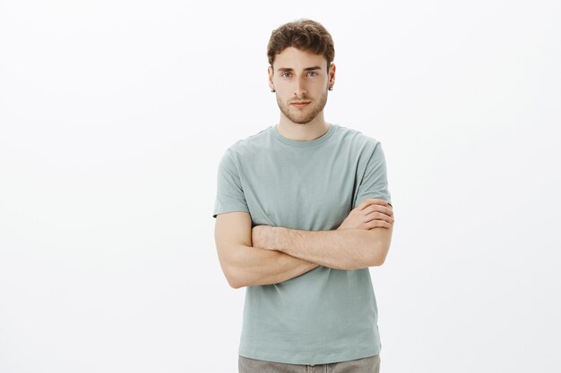 Indoor shot of confident good-looking european guy with fair hair in casual t-shirt, holding hands crossed on chest and looking with serious expression