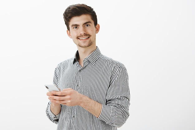 Indoor shot of confident joyful male model with moustache and beard, holding smartphone and smiling