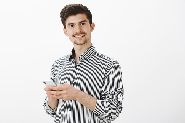 Indoor shot of confident joyful male model with moustache and beard, holding smartphone and smiling