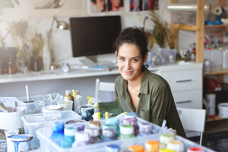 मुफ़्त फोटो indoor shot of creative female painter with beautiful appearance sitting at table surrounded with colorful oils, looking with glad expression