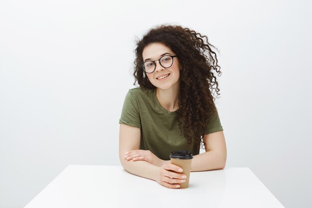 Indoor shot of creative good-looking female businesswoman with curly hair in eyewear, sitting at table and drinking coffee, smiling broadly