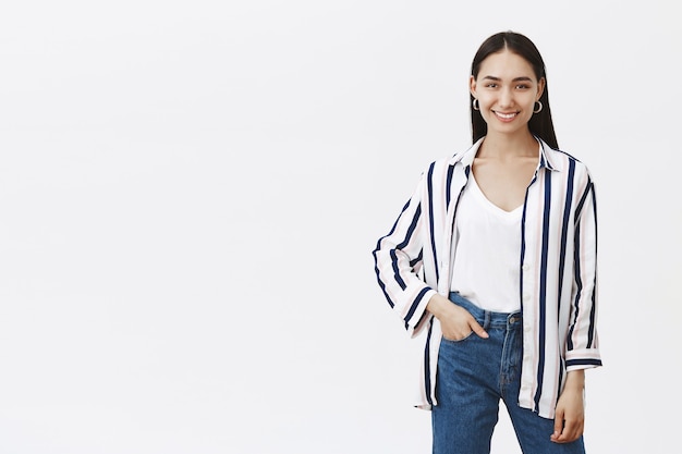 Indoor shot of creative good-looking stylish designer in striped blouse and stylish jeans, holding hand in pocket while smiling broadly, standing in relaxed and confident pose over gray wall