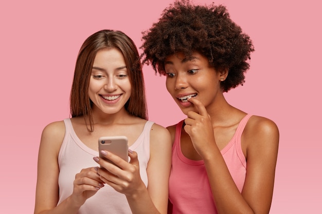 Indoor shot of curious cheerful rwo multiethnic women read news in internet via cell phone, make purchases online, stands closely, have interracial relationships, isolated over pink wall.