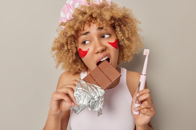 巻き毛の女性が歯に有害な甘いチョコレートを噛む屋内ショットは、電動歯ブラシが美容処置を受け、灰色のスタジオの背景に対して目のポーズの下に赤いパッチを適用します
