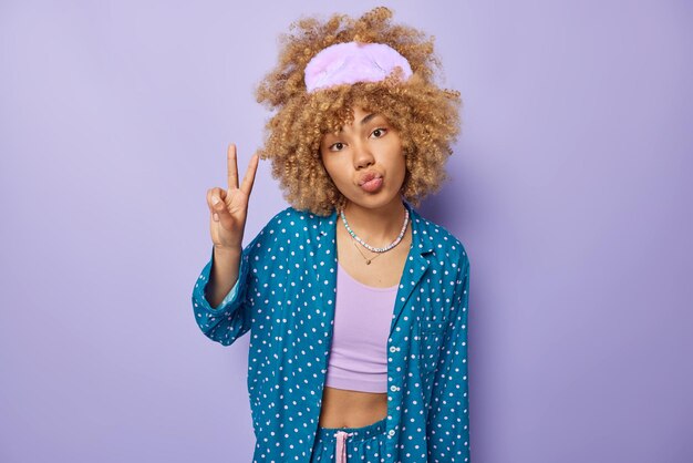 Indoor shot of curly haired woman wears sleepmask and comfortable pajama keeps lips folded gestures victory or peace sign isolated over purple background Good morning and awakening concept