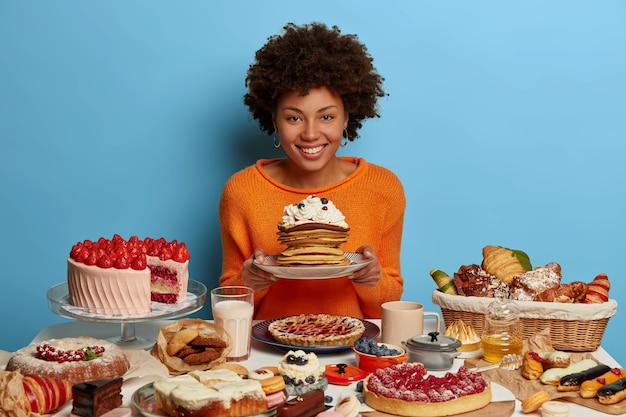 Foto gratuita tiro al coperto di donna riccia ha pranzo da forno dolce, tiene il piatto con deliziose frittelle cremose zuccherate, circondato da dolci al forno