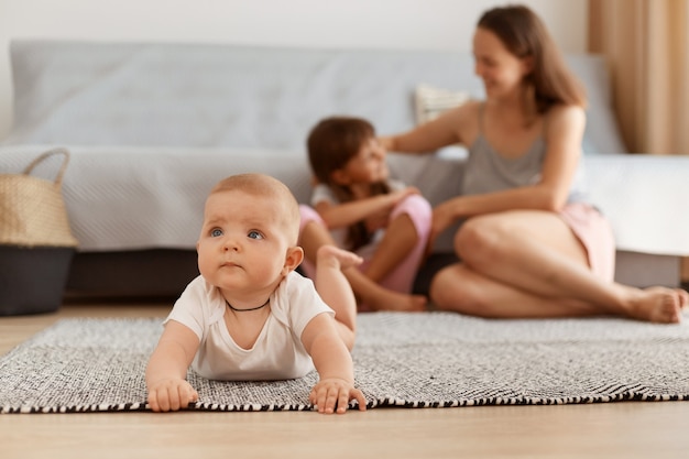 귀여운 아기 소녀가 카펫 바닥에 엎드려서 관심을 갖고 올려다보고 호기심 많은 표정을 하고 소파 근처에 앉아 배경에 앉아 있는 어머니와 여동생의 실내 사진.