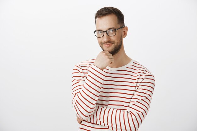 Indoor shot of cute adult man bartender in black glasses, touching beard and smiling curiously