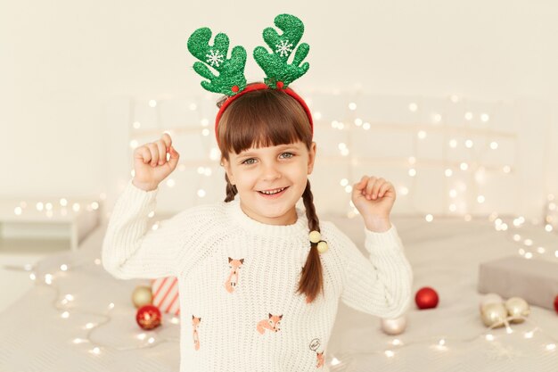 Indoor shot of cute charming kid girl wearing white sweater and party green deer sitting on bed with raised arms, looking at camera with smile, celebrating new year.