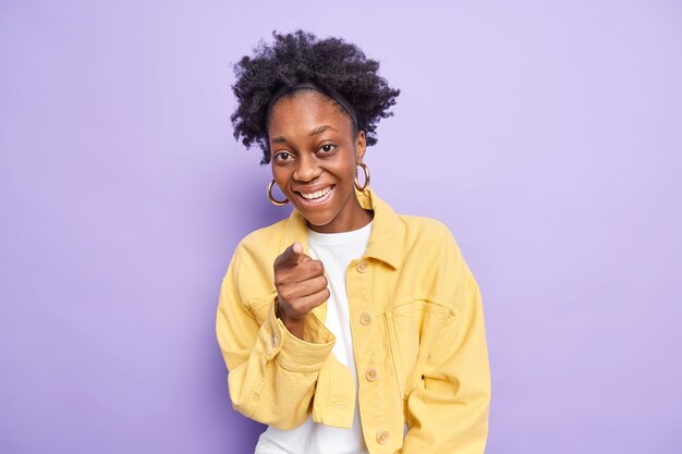 Indoor shot of cute dark skinned woman with curly hair indicates directly at camera