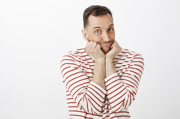 Indoor shot of cute emotive gay man in striped pullover, leaning face on palms and making tender duck face with pouting lips