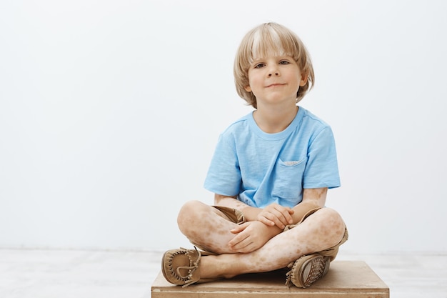 Tiro al coperto di carino felice bambino biondo con un sorriso positivo seduto con le mani incrociate, con vitiligine, sorridente largamente mentre appendere fuori con gli amici all'asilo