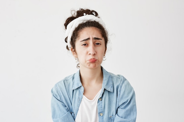 Indoor shot of dark-haired upset female being abused by someone, curves lips, frowns, looks unhappy, wears denim shirt, isolated against white wall. Negative human emotions and feelings