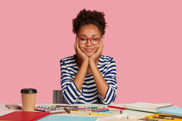 Free photo indoor shot of dark skinned fashion designer with crisp hair, touches cheeks with both hands, wears striped clothes, works on new creative project, poses at workplace against pink wall