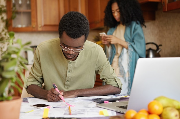 Indoor shot of dark-skinned man sitting at kitchen table using pink felt pen