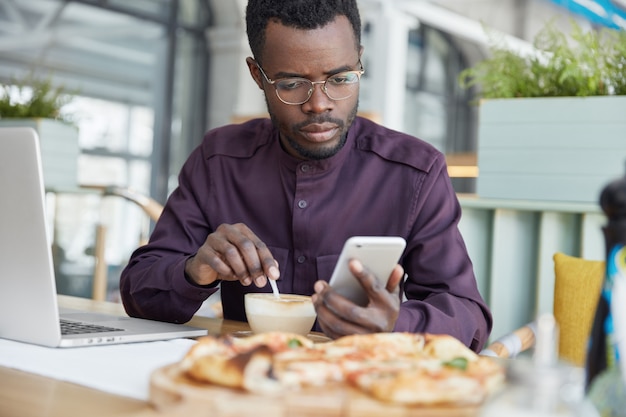 Indoor shot of dark skinned serious young African male enterpreneur focused into screen of mobile phone, drinks latte, reads attentively news on internet website