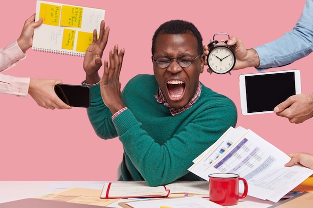 Indoor shot of desperate young Afro American man screams desperately, makes stop gesture, dressed in green sweater, busy working, isolated over pink background. People