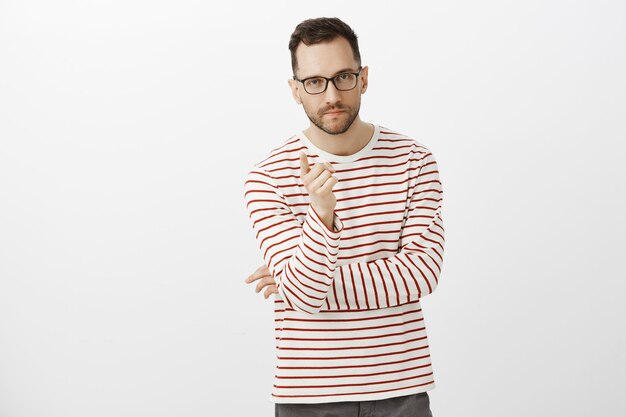 Indoor shot of determined creative businessman in black glasses and striped shirt, pointing with index finger and looking seriously, giving assignment to office team, standing over gray wall