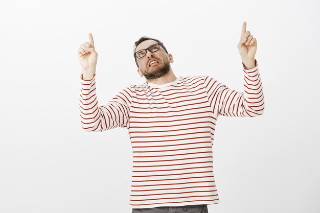 Indoor shot of displeased disgusted adult guy in fancy striped pullover, pointing and looking up with antipathy and dislike