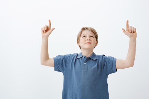 Indoor shot of displeased doubtful man with blond hair, looking and pointing up with raised index fingers, frowning, being unsure and annoyed with loud neighbours