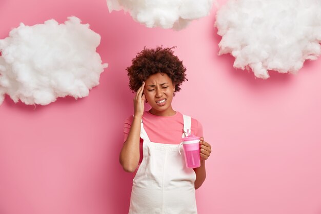 Indoor shot of displeased pregnant woman suffers from headache, feels unbearable migraine, has big tummy, drinks water, dressed in casual clothes, isolated on pink wall. Family and pregnancy