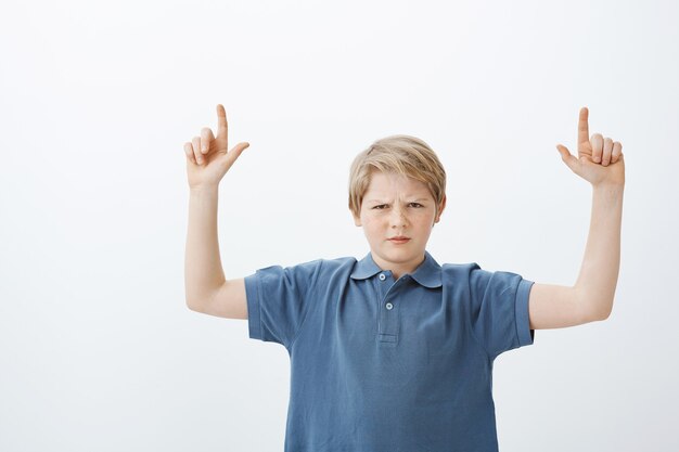 Снимок недовольной несчастной блондинки в помещении: ребенок в синей футболке хмурится и поднимает указательные пальцы, указывая вверх