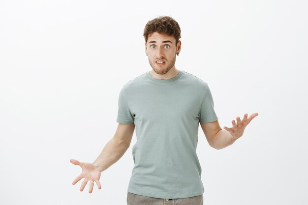 Indoor shot of displeased upset attractive guy in casual t-shirt, gesturing with spread palms and staring