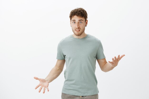 Внутренний снимок недовольного расстроенного привлекательного парня в повседневной футболке, жестикулирующего ладонями и смотрящего