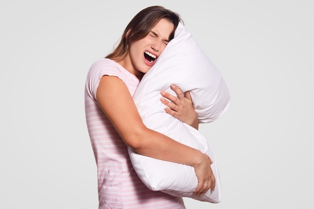 Indoor shot of displeased woman opens mouth widely, has dissatisfied expression, keeps white pillow in hands, wears casual domestic clothes, models indoor, frustrated to have horrified dreams at night